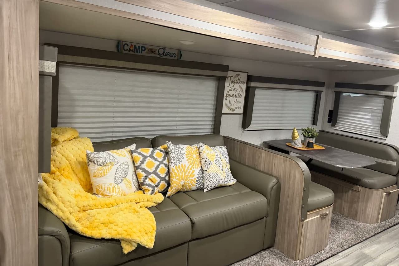 Inside the trailer: a long couch under the windows with yellow cushions and a throw, and the dinette beyond it.